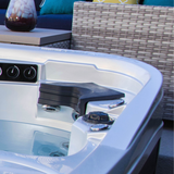 A charcoal black Artesian Elite neck headrest pillow installed in the corner seat of a luxury white acrylic hot tub, positioned over the integrated spa jets with outdoor patio furniture in the background.