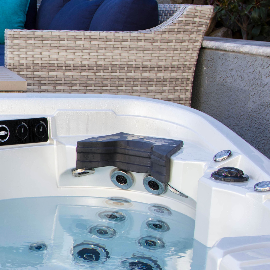Close-up view of a charcoal black Artesian Elite neck headrest pillow installed in a white acrylic hot tub seat, positioned directly above three chrome hydrotherapy jets with clear water in the foreground.