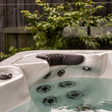 An Artesian Spas Island series neck collar charcoal grey headrest pillow installed in an outdoor backyard hot tub above a hydrotherapy jet cluster.