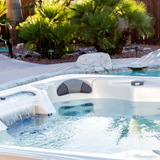 An Artesian Spas Island Lounge charcoal grey headrest pillow installed in an outdoor backyard hot tub next to a running waterfall feature and surrounded by palm trees.