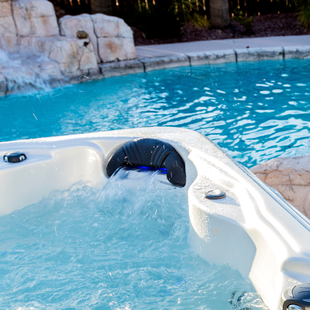 An Artesian Spas Island series charcoal grey waterfall headrest pillow with active blue LED lighting illuminating the water flow, installed in an outdoor hot tub next to a swimming pool.