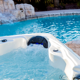 An Artesian Spas Island series charcoal grey waterfall headrest pillow with active blue LED lighting illuminating the water flow, installed in an outdoor hot tub next to a swimming pool.
