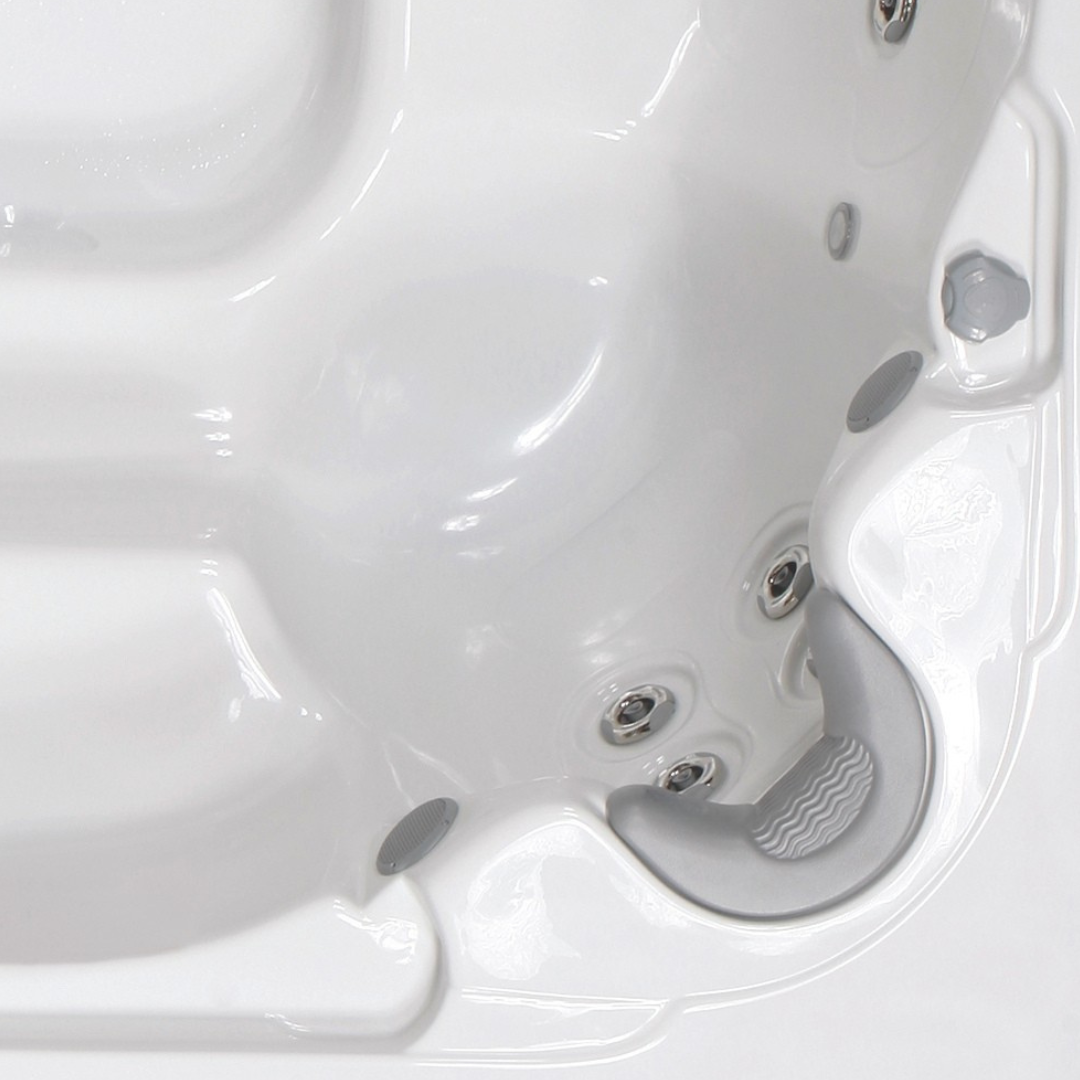 Overhead view of a curved silver grey foam neck headrest pillow without a logo, installed in a white acrylic hot tub corner seat near water jets and air dials.