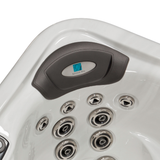 Top-down angle view of a grey curved replacement foam headrest pillow with its central silver logo dome installed in a white acrylic hot tub corner seat, located above a cluster of chrome massage jets.