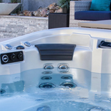A charcoal black curved Elite corner headrest pillow installed in an outdoor hot tub filled with clear blue water, positioned above a vertical row of massage jets with patio furniture and a stone wall in the background.