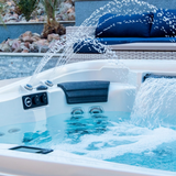 A charcoal black curved Elite corner headrest pillow installed in a running outdoor hot tub, framed by arching water fountain streams and a cascading sheet waterfall feature with patio seating in the background.