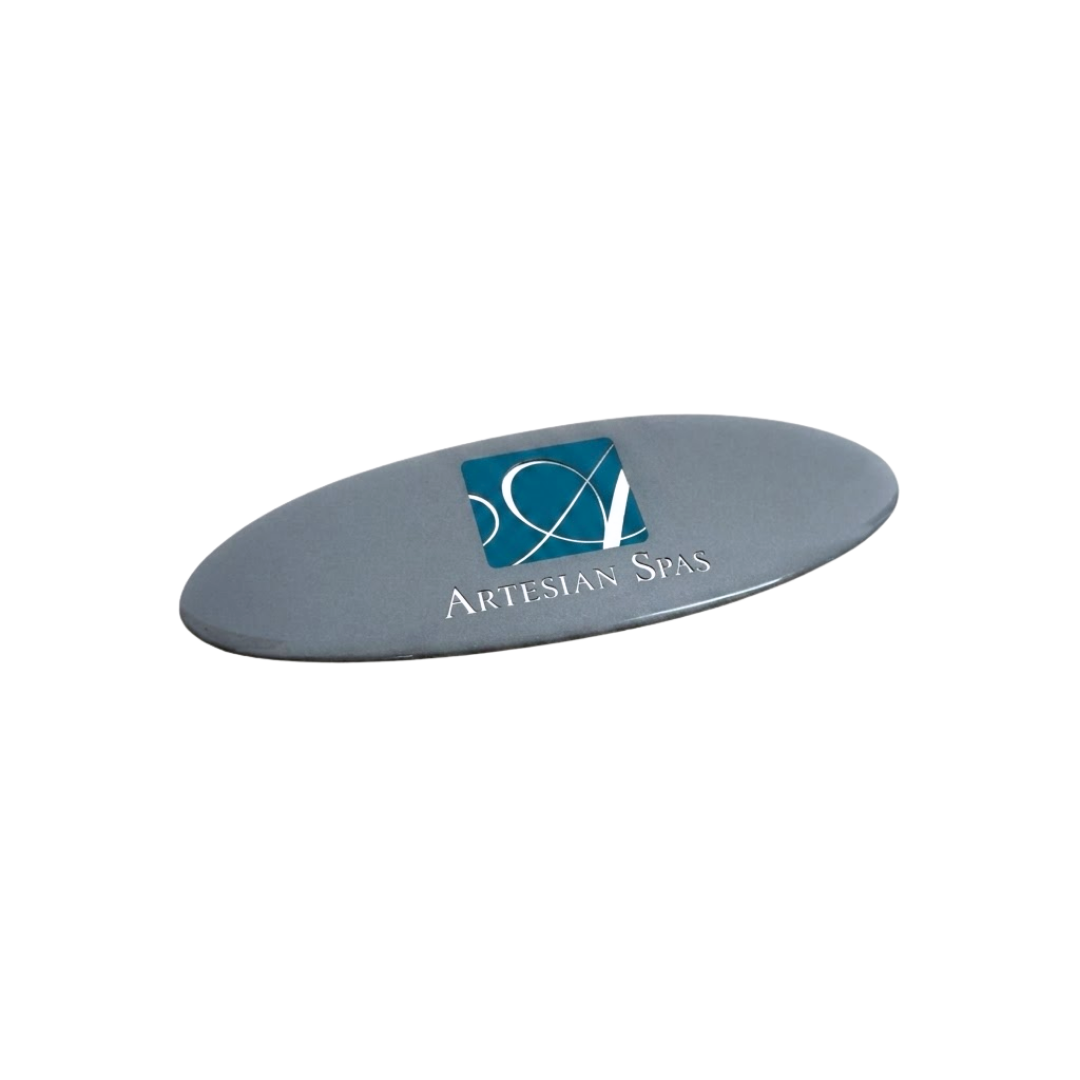Angled perspective view of an oval metallic silver replacement resin dome emblem for an Artesian Spas headrest pillow, featuring the teal square cursive "A" logo and "ARTESIAN SPAS" text.