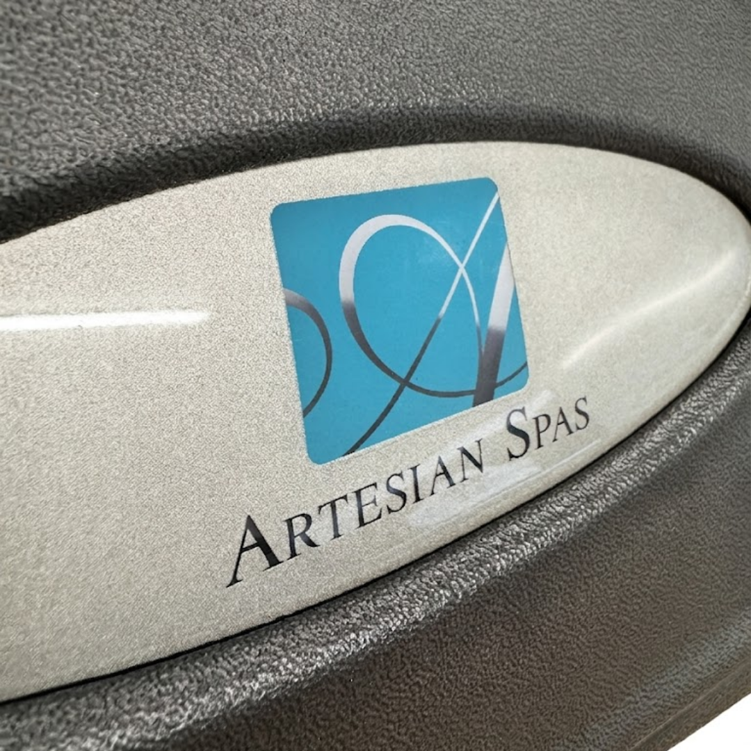 Macro close-up view of an oval metallic silver resin dome logo badge inserted into a grey textured foam replacement headrest pillow for an Artesian Spas hot tub, showcasing the glossy finish, teal branding square, and crisp typography.