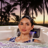 A woman relaxing in an Artesian Spas Island series hot tub with water cascading over her neck from an 8-inch charcoal waterfall headrest pillow, set against a tropical sunset background with palm trees.