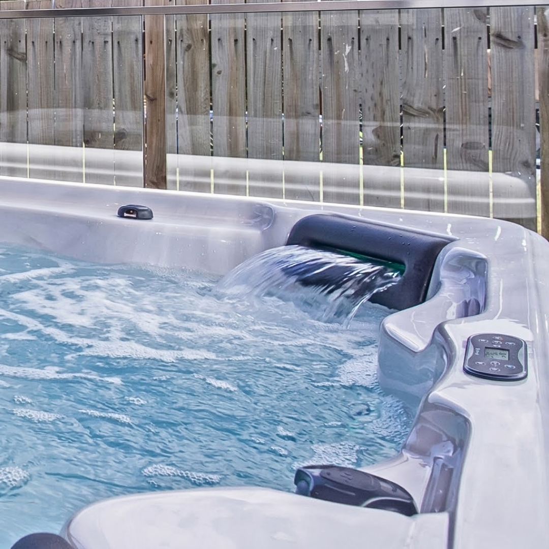 Artesian Spas Island series 8-inch waterfall headrest pillow in charcoal gray flowing water into an outdoor hot tub situated near a wooden fence and glass balustrade.