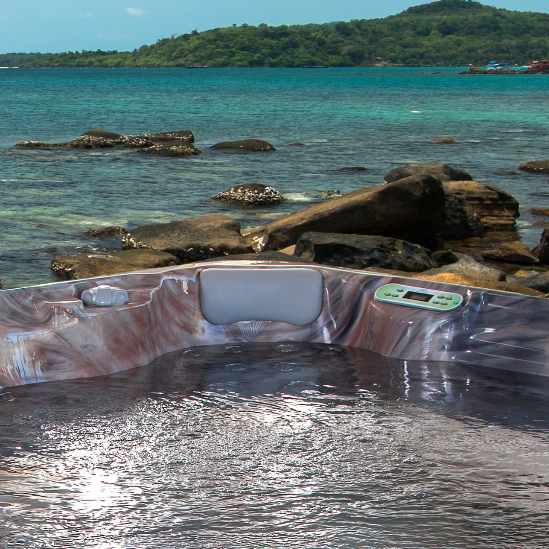 Artesian Spas South Seas Lounge series light gray headrest pillow installed in a marbled brown acrylic hot tub corner, set against a scenic tropical ocean backdrop.