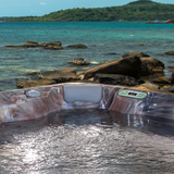 Artesian Spas South Seas Lounge series light gray headrest pillow installed in a marbled brown acrylic hot tub corner, set against a scenic tropical ocean backdrop.