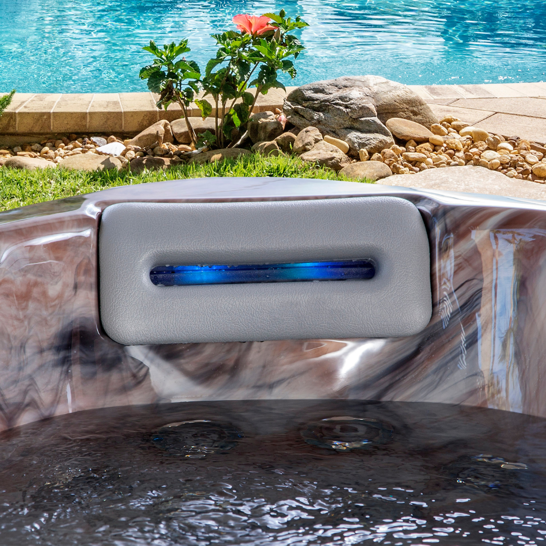 Artesian Spas South Seas and Tropic Seas series 8-inch waterfall headrest pillow in light gray with integrated blue LED lighting, installed in a marbled hot tub near a poolside garden.