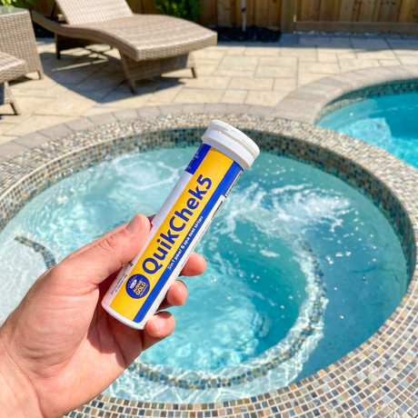 A hand holding a bottle of Aussie Gold QuikChek5 5-in-1 test strips over a backyard pool and spa, used for quick DIY water chemistry analysis.