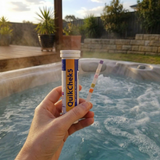 A hand holding an Aussie Gold QuikChek5 test strip showing color results next to the bottle, held over a bubbling outdoor hot tub for an instant water chemistry check.