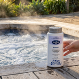 A bottle of BioGuard SPA Armour sanitiser and oxidiser sitting on a weathered wooden pool deck next to a steaming, bubbling outdoor spa. This chlorine and bromine dual-action formula is designed for bacteria and algae control in outdoor spa environments.