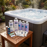 An assortment of BioGuard Spa water treatments testing strips and maintenance chemicals arranged on a wooden table next to a steaming outdoor hot tub