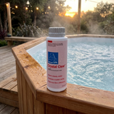 A bottle of BioGuard SPA Crystal Clear water clarifier balanced on the wet wooden edge of an outdoor hot tub at sunset. The pink label indicates the formula clears cloudy water, restores crystal clarity, and improves filter efficiency for backyard spas and jacuzzis.