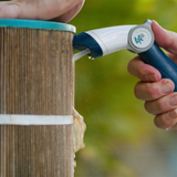 A close-up action shot of a person using the handheld Life Water-Wand Pro sprayer to blast a high-pressure stream of water into the pleats of a dirty, brown hot tub cartridge filter, washing away trapped debris against a blurred green background.