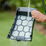 A person rinsing the black and white honeycomb filter basket tray of a Maytronics Dolphin Skimmi 300 solar pool cleaner with a garden hose.