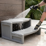 A person wearing utility gloves sprays water from a garden hose nozzle to clean a grey plastic two-tier Confer Plastics Handi-Step hot tub entry stair unit.