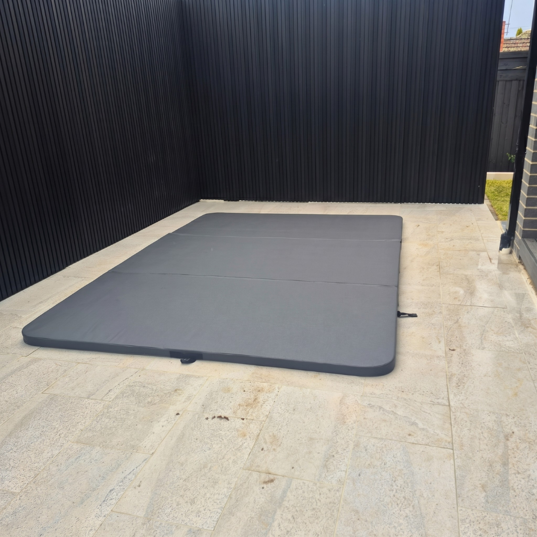 A large custom dark grey three-panel hard cover installed over an inground swim spa on a light stone tiled patio with a modern black slatted fence background.