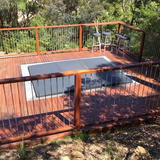 A large custom grey hard cover fitted over an inground spa built into an elevated wooden timber deck with a stainless steel wire railing.