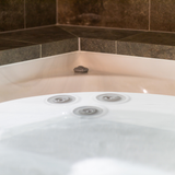 Close-up view of grey Cyclone adjustable swirl spa jets installed in the white wall of an indoor hot tub adjacent to dark stone tiled decking.