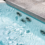 Angled shot of submerged grey Cyclone adjustable swirl spa jets operating along the wall of a clear blue swimming pool below granite coping stone.