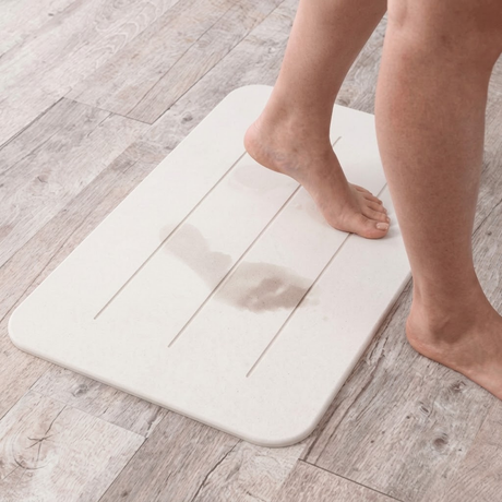 Action photo of bare feet stepping onto a white Drysorb diatomaceous earth bath mat with grooved lines, demonstrating fast moisture absorption leaving damp footprints on a light wood-look floor.