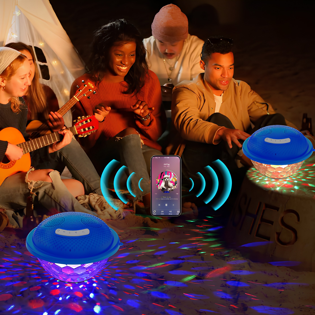 Group of friends sitting around a campfire on a beach pairing a smartphone to two floating Bluetooth pool speakers with multicolored lights for stereo sound.