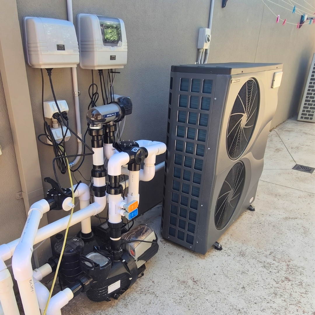 An installed tall, dark grey EvoHeat Force iPro series inverter pool heat pump featuring two stacked front-discharge fan grilles, positioned alongside its connected white PVC plumbing pipes, water pumps, and wall-mounted control boxes on a concrete patio against a residential wall.