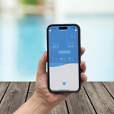 Close-up view of a hand holding a smartphone displaying a pool heat pump mobile application interface with temperature readings, set against a blurry outdoor swimming pool background.