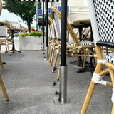 Line of heavy-duty stainless steel EzyHold commercial umbrella holder sleeves bolted flush into an asphalt sidewalk cafe patio with black bistro chairs and tables