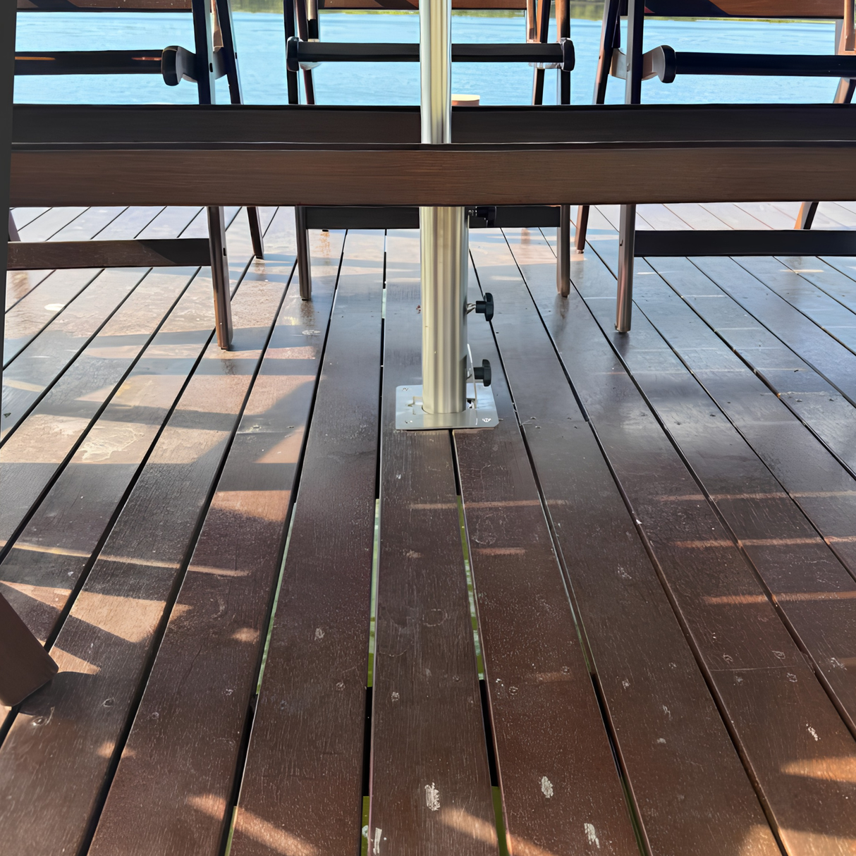Heavy-duty stainless steel EzyHold umbrella holder sleeve bolted flush onto a dark timber boardwalk boat dock overlooking open blue water.