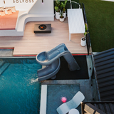 High angle view of an S.R.Smith Helix2 grey spiral water slide running into an inground swimming pool next to a modern timber deck with a built-in fire pit