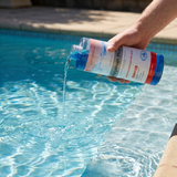 A person pouring blue liquid from a bottle of BioGuard Polysheen Plus super concentrated pool water clarifier directly into a sunny backyard swimming pool to clear up cloudy water and improve filtration.