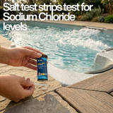 A person holding a bottle of Hy-Clor salt test strips and a test strip by an outdoor swimming pool with large white overlay text reading Salt test strips test for Sodium Chloride levels