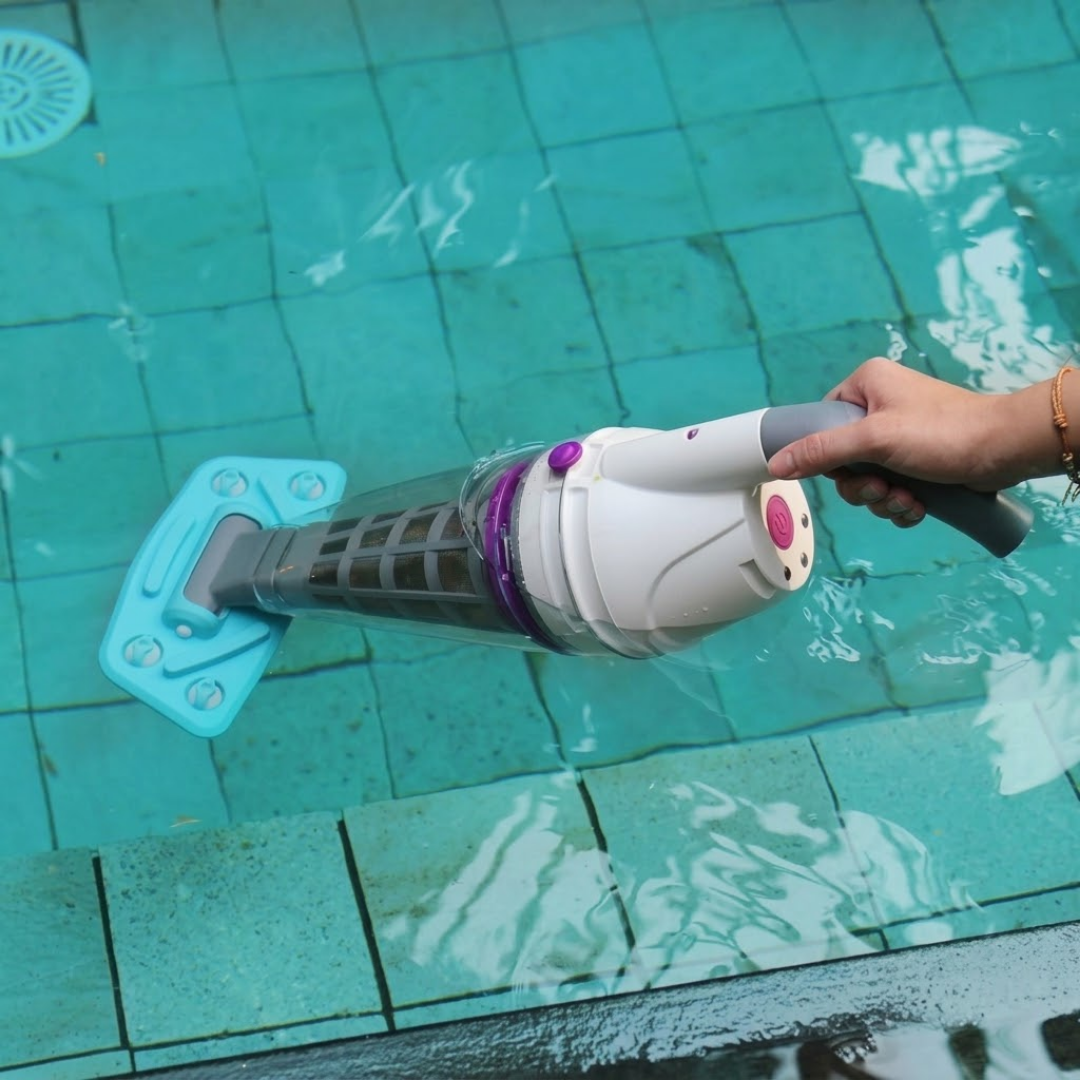 Action photo of a person holding the handle of a white and blue Kokido Telsa 50 cordless pool vacuum partially submerged beneath clear water along the edge of a tiled swimming pool.