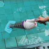 Action photo of a person holding the handle of a white and blue Kokido Telsa 50 cordless pool vacuum partially submerged beneath clear water along the edge of a tiled swimming pool.