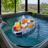 An inflatable silver Life Spa Bar snack tray and beverage holder floats in the clear water of a large indoor hot tub. The bar is stocked with a bottle of wine on ice, canned drinks, tortilla chips with dip, and fresh fruit, set against a sunroom background overlooking a lush green valley and distant city skyline.