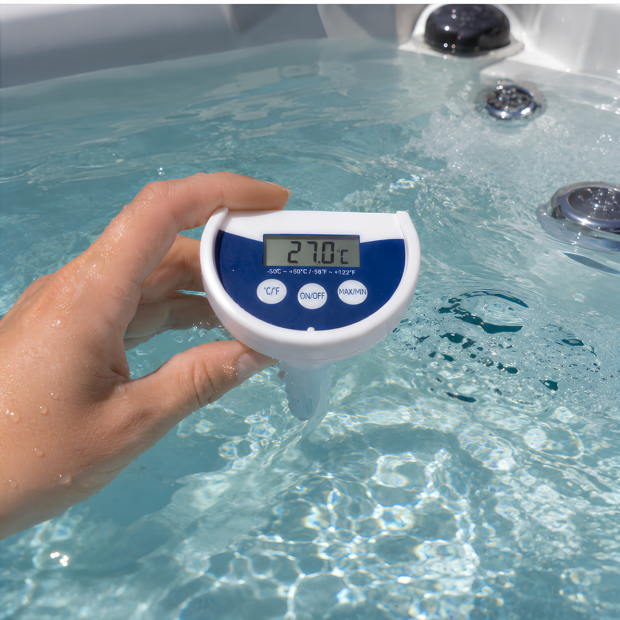 A close-up lifestyle shot of a hand holding a white and blue Life Spa Essentials floating digital thermometer in a hot tub. The LCD screen displays a clear water temperature reading of 27.0°C, with the hot tub jets bubbling in the background.