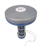 An elevated, top-down studio product shot of a gray and blue Life Spa Essentials floating hot tub chemical dispenser, isolated on a white background. The image highlights the round gray floating cap with embossed warnings and a central white Life logo, looking down onto the slotted gray basket and a dangling white anchor string.