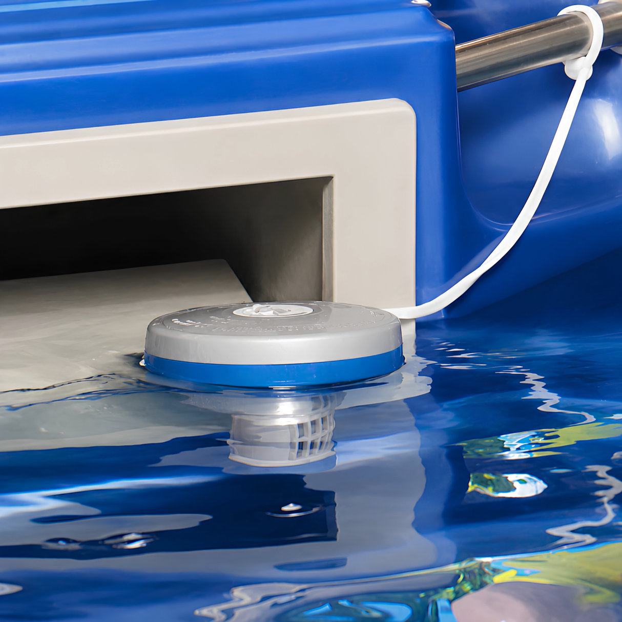 A close-up lifestyle shot of a gray and blue Life Spa Essentials floating chemical dispenser bobbing on clear blue pool water. A white anchor cord tethers the floating chlorinator to a silver metal handrail on a blue pool structure, with the submerged slotted tablet basket visible just below the surface.
