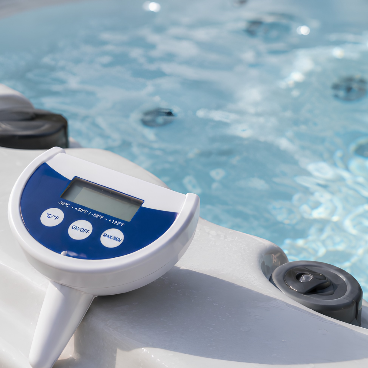 A close-up lifestyle shot of a white and blue Life Spa Essentials floating digital thermometer sitting on the gray acrylic ledge of an outdoor hot tub, highlighting its clear LCD display screen, round control buttons, and the bubbling blue spa water in the background.