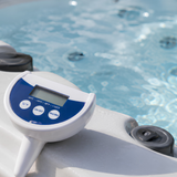 A close-up lifestyle shot of a white and blue Life Spa Essentials floating digital thermometer sitting on the gray acrylic ledge of an outdoor hot tub, highlighting its clear LCD display screen, round control buttons, and the bubbling blue spa water in the background.