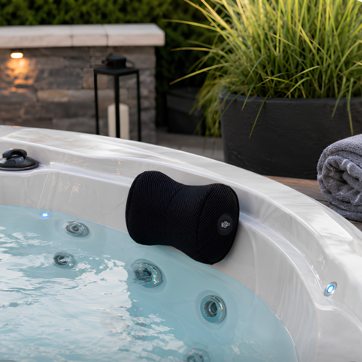 A contoured black Life Spa Pillow headrest mounted on the edge of a white luxury outdoor hot tub with bubbling water and glowing blue LED lights, featuring a rolled gray towel, a large potted grass plant, and a stone wall accent in the background.