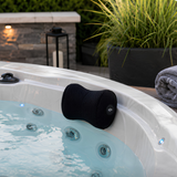 A contoured black Life Spa Pillow headrest mounted on the edge of a white luxury outdoor hot tub with bubbling water and glowing blue LED lights, featuring a rolled gray towel, a large potted grass plant, and a stone wall accent in the background.