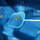 Overhead action photo of a light blue spa skimmer net attached to a telescopic pole, collecting floating leaves from the clear water inside a blue acrylic hot tub.