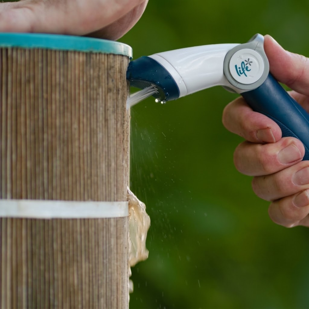 Action photo of a person holding the Life Spa Water-Wand Pro sprayer to flush trapped debris and dirt from the pleats of a dirty pool or spa cartridge filter, set against a soft blurred green outdoor background.